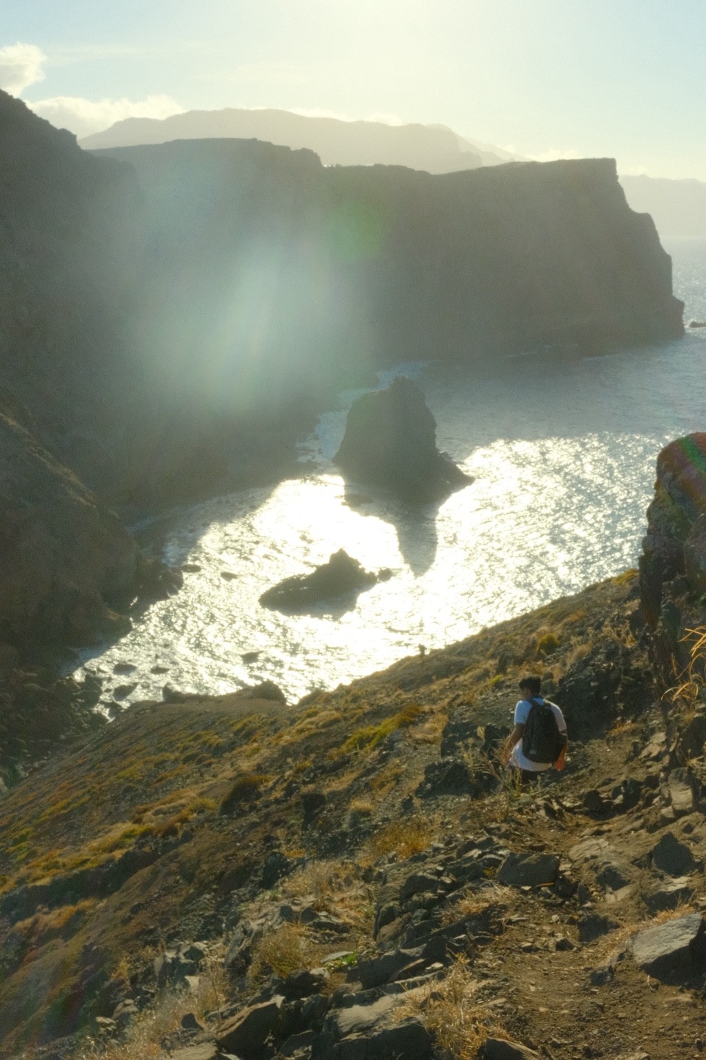 Sunlight glints off the sea between layered cliffs, with a small hiker on the rocky slope.